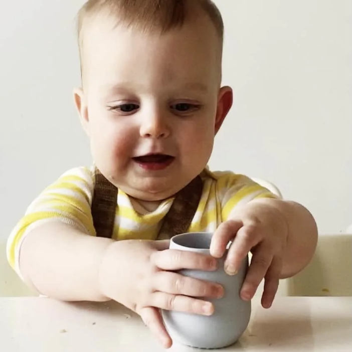 Baby holding gray silicone cup, mess-free mealtime with easy clean food catcher mat
