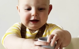 Baby holding gray silicone cup, mess-free mealtime with easy clean food catcher mat