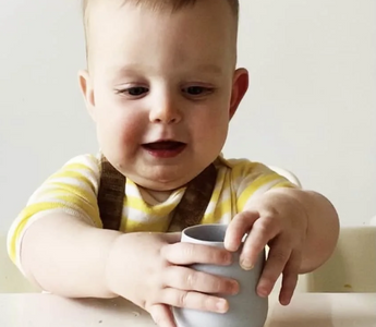 Baby holding gray silicone cup, mess-free mealtime with easy clean food catcher mat
