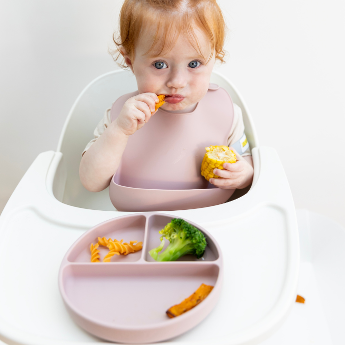 Baby eating corn and pasta with mess-free silicone bib and divided plate, easy clean, food-safe design
