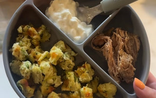 Gray divided baby plate with broccoli bites, yogurt, and shredded meat, held by hand with orange nail polish.