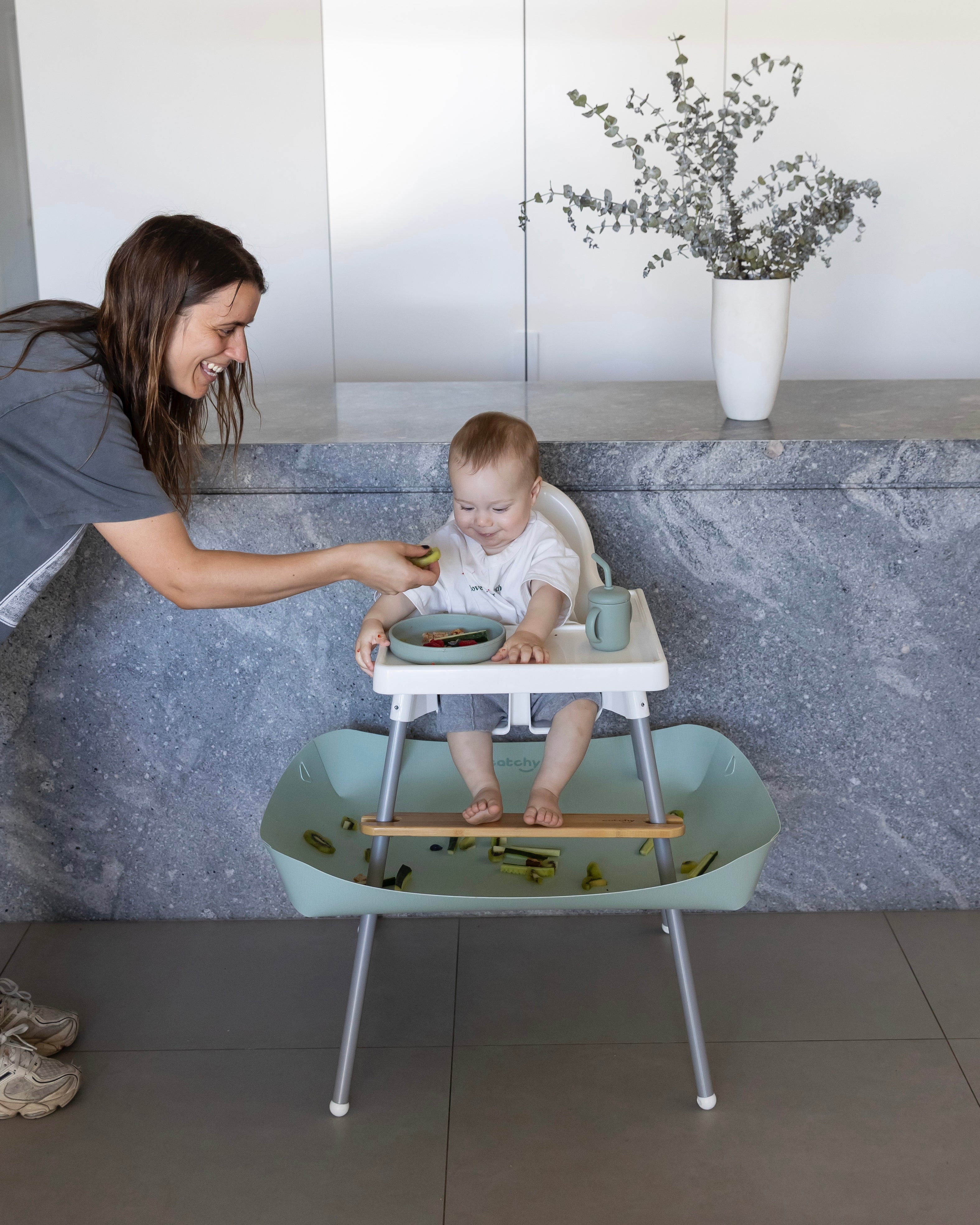 Baby in high chair with mess-free food catcher, mom feeding with smile